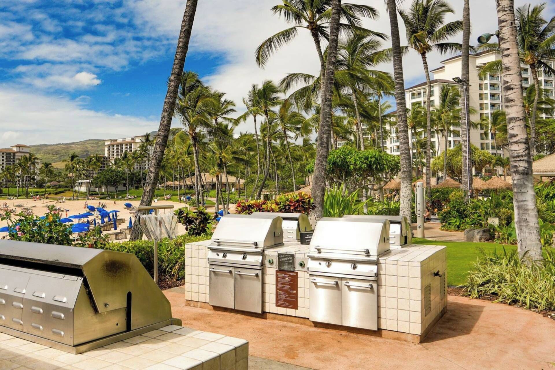 Ein Grillbereich im Freien mit mehreren Grillstationen, umgeben von Palmen und mit Blick auf ein Hotelgebäude im Hintergrund.