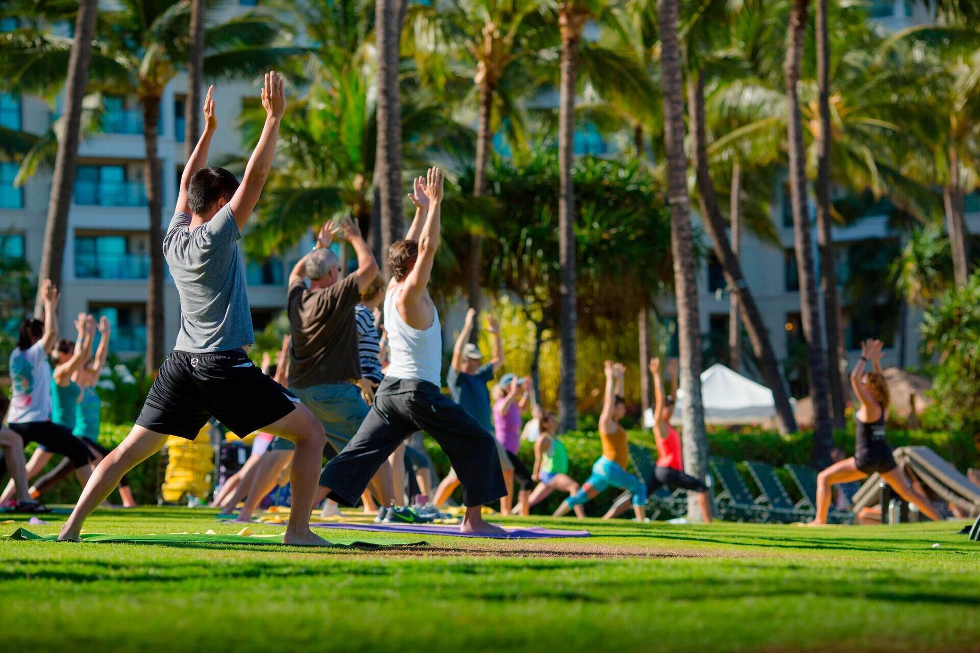 Un groupe de personnes pratiquant un exercice de yoga en plein air, avec des palmiers et un bâtiment en arrière-plan.