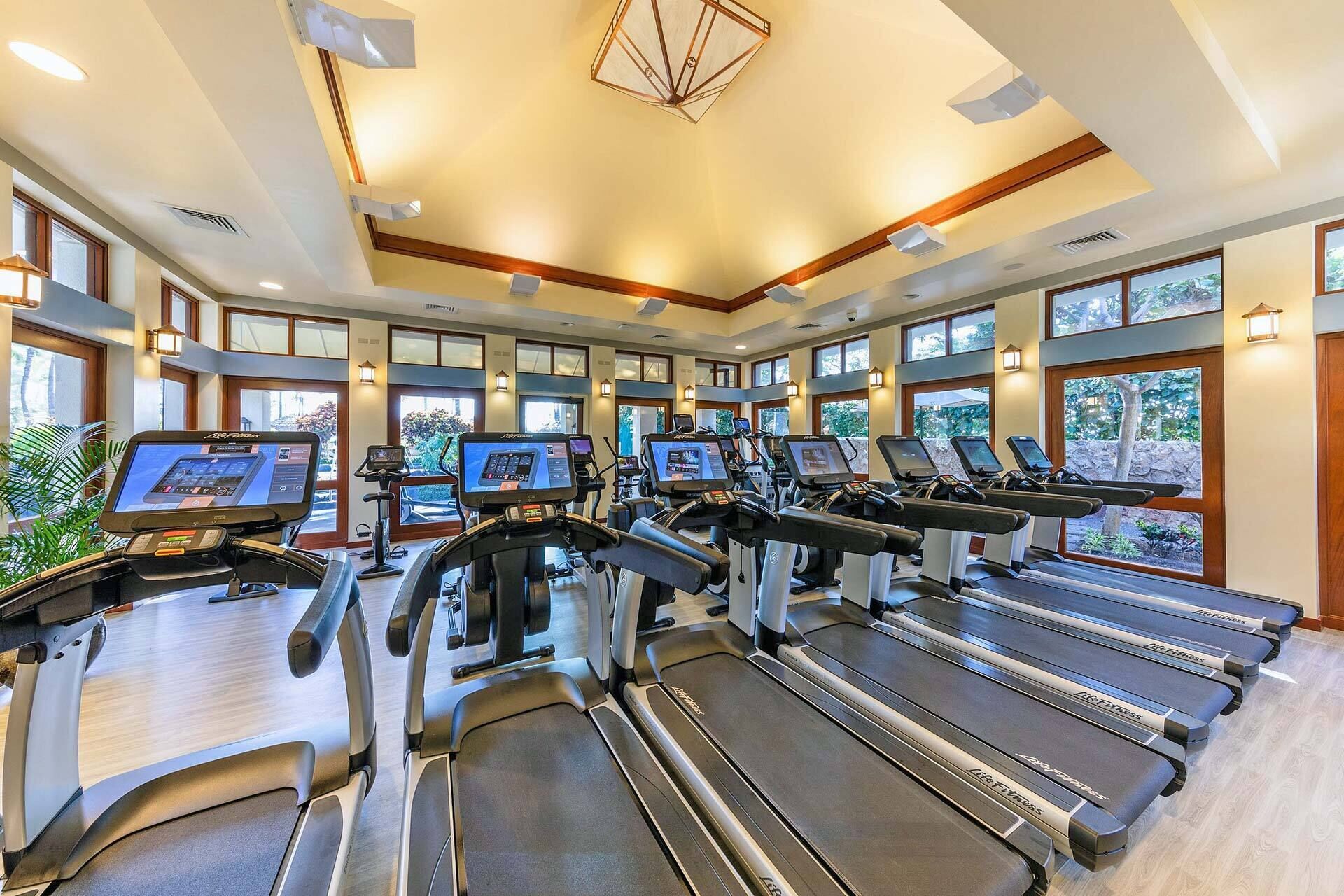 Un gimnasio con cintas de correr, bicicletas estáticas y elípticas, amplias ventanas y un diseño de techo moderno.