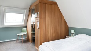 Een slaapkamer met een schuine kap, een houten garderobe, een groene stoel, een bed en een nachtkastje met een lamp.