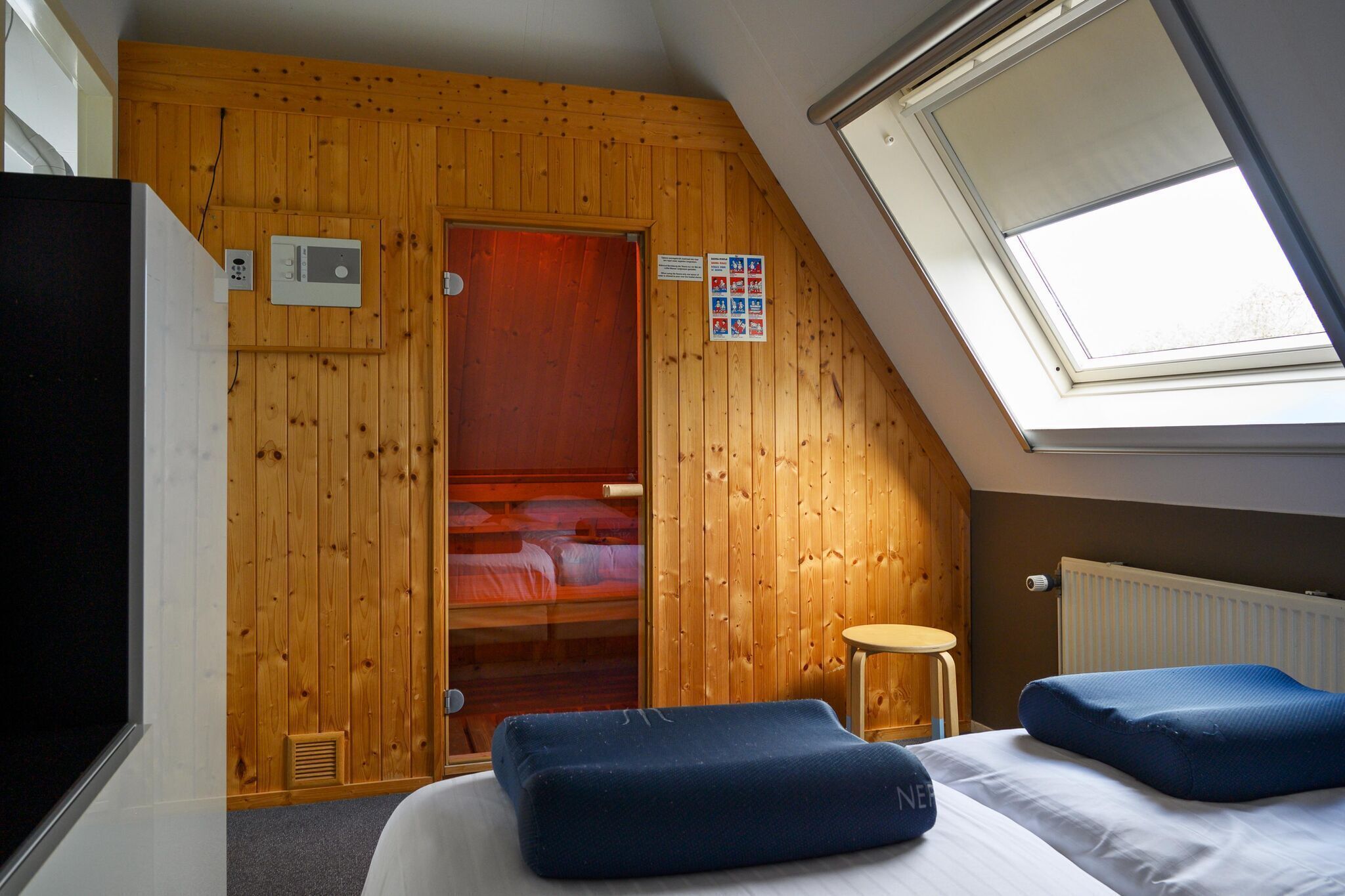 A room with wooden walls, a skylight, a bed with blue pillows, a small stool, and a radiator.
