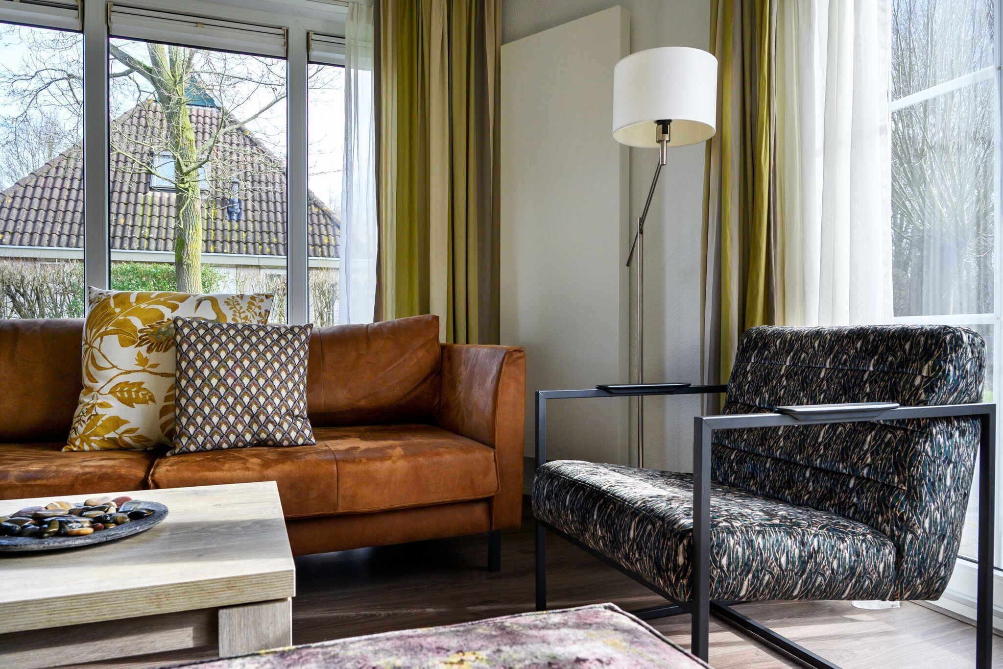 A modern living room with a brown leather sofa, patterned cushions, a patterned armchair, a side table, and a floor lamp.