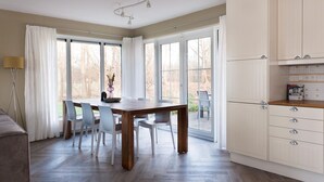 Ein Essbereich mit Holztisch und Stühlen, eine Küche mit weißen Schränken und große Fenster mit Blick auf Bäume.