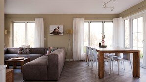 Ein Wohnzimmer mit einer Couch, einem Esstisch mit Stühlen und großen Fenstern mit Vorhängen.