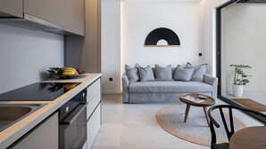 A modern kitchen with a built-in oven, a dining area with a round table and chairs, a grey sofa, and a large window with a view of the outdoors. - Urban Oasis - Walking distance to the Old Town, the beach and all kind of shops! (Chania)