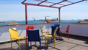 Une terrasse offrant une vue sur la mer et les collines, avec des chaises rouges, jaunes et bleues, ainsi qu’une table.
