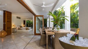 A spacious living area with a large dining table, wicker furniture, and a view of lush greenery outside. - Frangipani Villa | Port Douglas (PORT DOUGLAS)