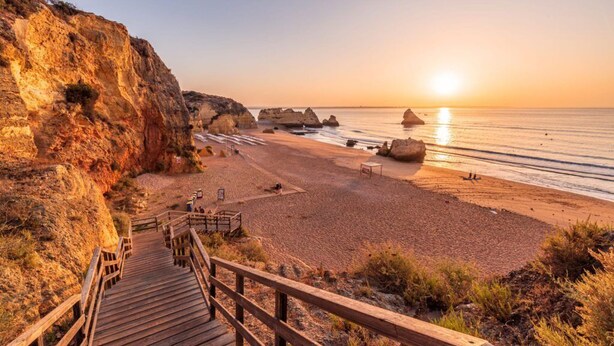 Una playa costera con una escalera de madera que desciende hacia la orilla, rodeada de acantilados rocosos y un cielo despejado.