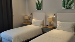 Een hotelkamer met twee bedden, elk voorzien van nachtkastje en lamp, en twee aan de muur bevestigde schilderijen van bladeren.