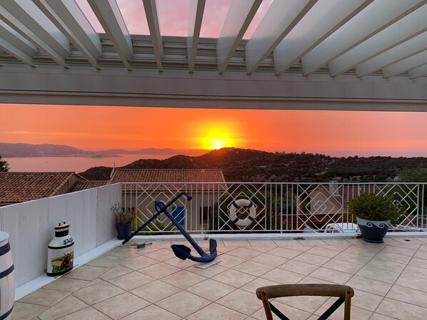 A balcony with a view of a sunset over a coastal landscape, featuring a railing, potted plants, and a chair.