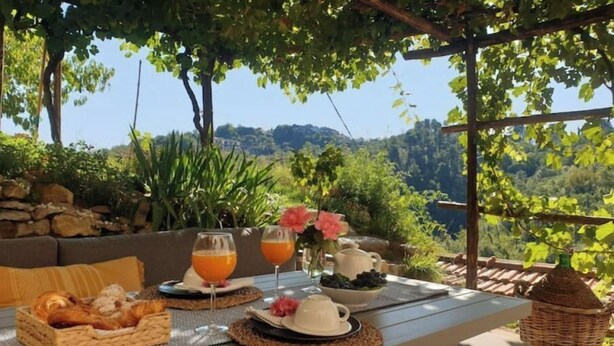 Ein Essbereich im Freien mit einem für das Frühstück gedeckten Tisch, darunter Croissants, Obst und Getränke, mit Blick auf eine malerische Landschaft.