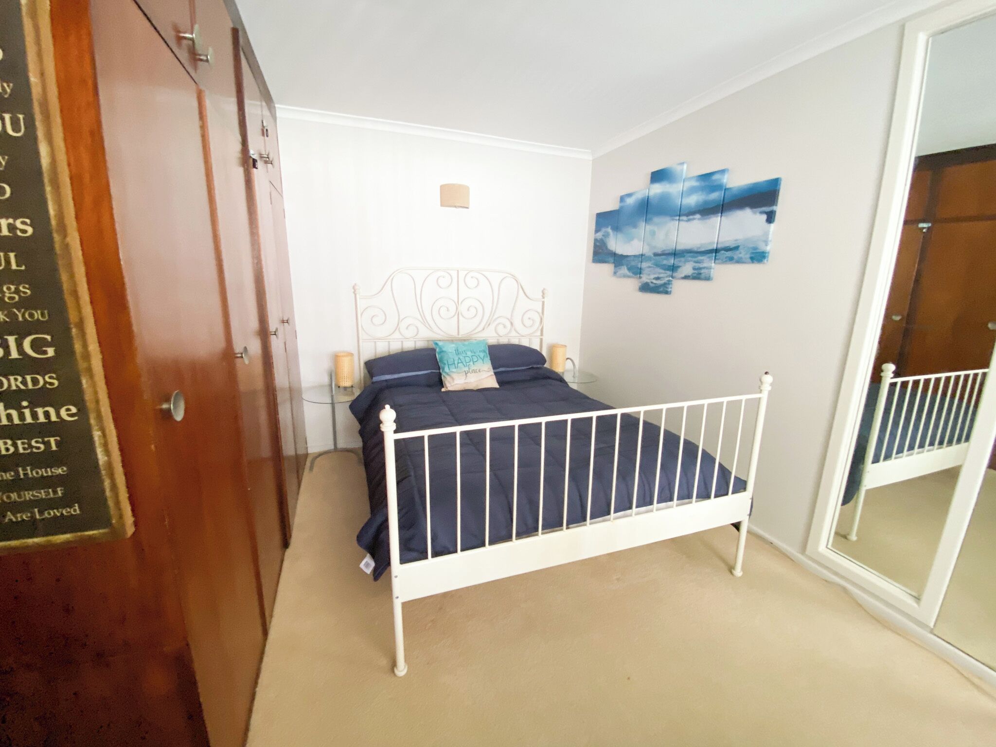 Een slaapkamer met een bed met wit metalen frame, een beigekleurig tapijt, een muur met een schilderij van een blauwe lucht, en een houten garderobe.