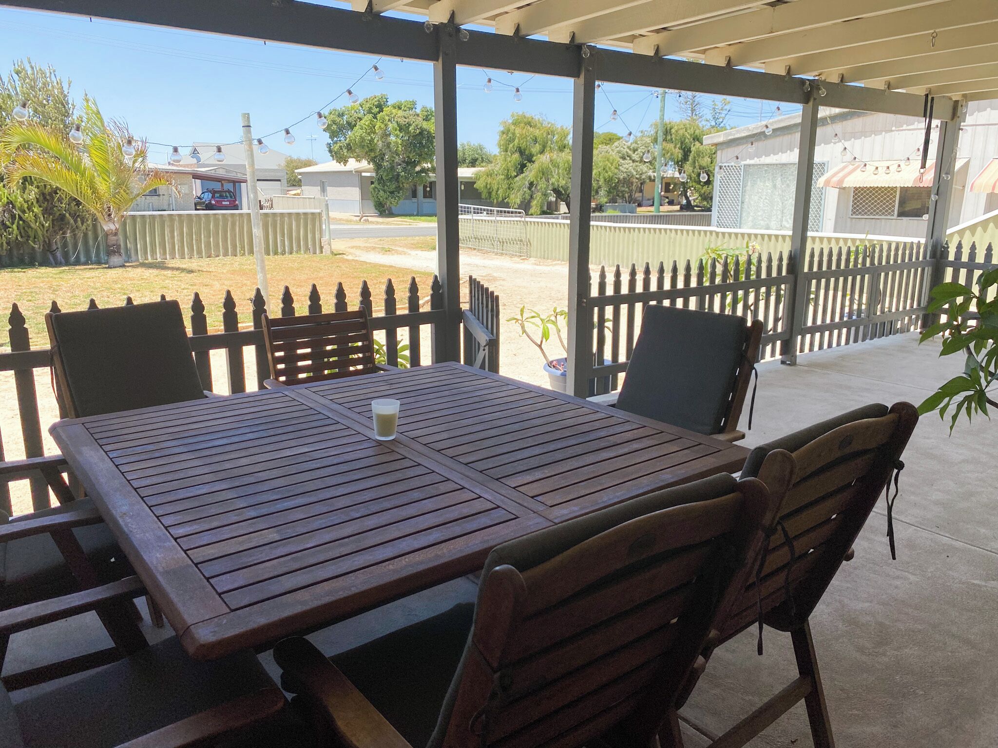 Une terrasse extérieure couverte, avec une table et des chaises en bois, un pot de fleurs et une vue sur le quartier.