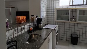 A kitchen with a granite countertop, stainless steel sink, and white tiled walls. There is a window with blinds, a black trash bin, and a small shelf with a kettle. - Praia de Manaíra (João Pessoa)