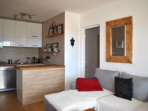 Eine moderne Küche mit weißen Schränken, einer Holz Arbeitsplatte und einem eingebauten Backofen. Ein graues Sofa mit einem roten und einem schwarzen Kissen mit Schneeflockenmuster. Ein spiegel mit Holzrahmen hängt an der Wand.