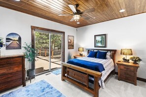 A bedroom with a wooden ceiling, a bed with a blue bedspread, a nightstand with a lamp, and a view of a deck through glass doors.