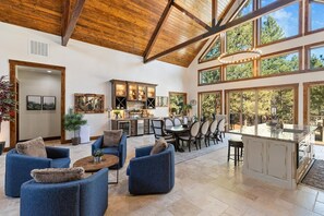 A spacious living area with a kitchen and dining area, featuring wooden beams, large windows, and modern furniture.