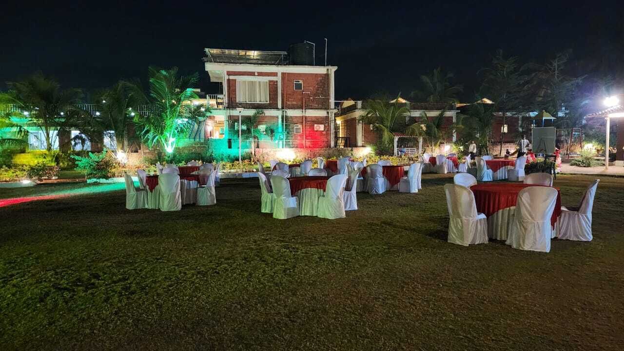 An outdoor event space with tables and chairs arranged for an event, a building in the background, and illuminated plants.