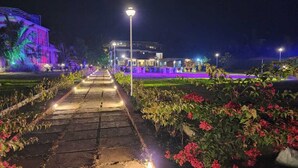 Un chemin menant à un bâtiment dont l’entrée est bien éclairée et qui est agrémenté d’un jardin où poussent des fleurs roses.