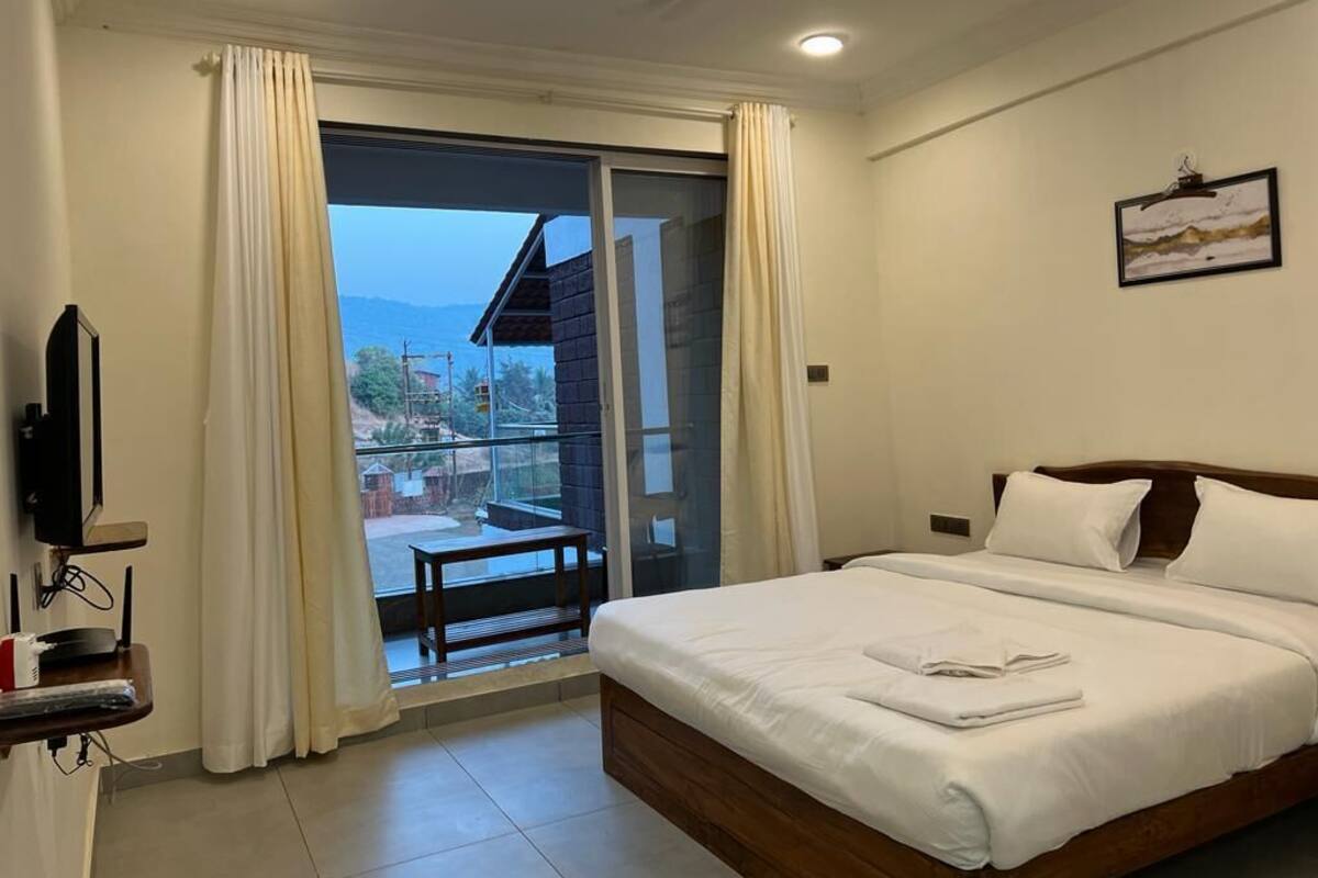 Een hotelkamer met een bed, een televisie, een balkon met gordijnen en uitzicht op gebouwen en bergen.