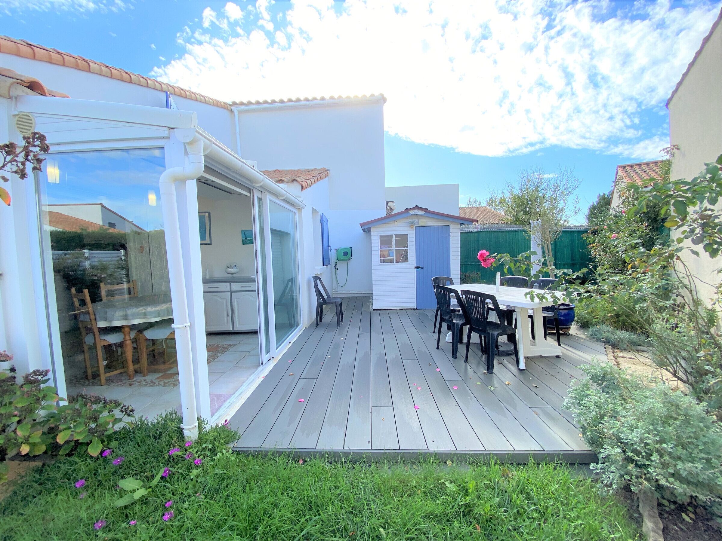 Une terrasse extérieure avec une table et des chaises, un petit abri de jardin et un espace clos par des parois de verre, à l’intérieur duquel on peut apercevoir une cuisine.