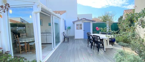 An outdoor patio with a dining table and chairs, a small shed, and a glass-enclosed area with a kitchen visible inside.