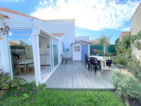 An outdoor patio with a dining table and chairs, a small shed, and a glass-enclosed area with a kitchen visible inside.