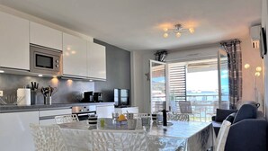 A modern kitchen with white cabinets, a microwave, and a coffee machine. There is a dining table with white chairs and a view of the outside through large windows. - Modern Apartment with Pool, Terrace, and A/C Near Beach - Le Lavandou (Le Lavandou)