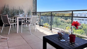 A balcony with a table and chairs, a red flower vase, and a view of trees and buildings. - Modern Apartment with Pool, Terrace, and A/C Near Beach - Le Lavandou (Le Lavandou)