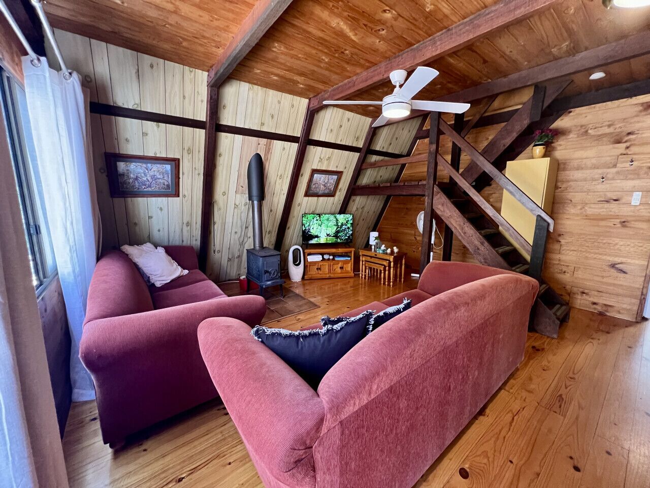 Acogedor espacio tipo loft con paredes de madera y techo inclinado, que presenta un sofá rojo, un mueble de entretenimiento de madera y un ventilador de techo.