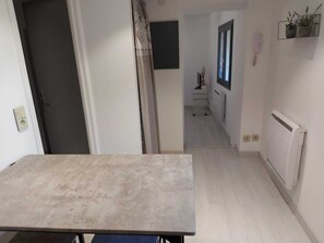 En smal gang med et bord i forgrunden, en dør til venstre, en radiator til højre og et vindue med en plante på væggen.