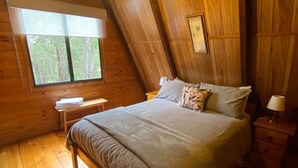 Een houten slaapkamer met een groot bed, een nachtkastje, een bankje en een raam met uitzicht op een bos.