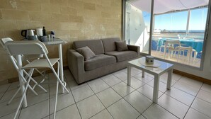 A room with a sofa, a small table, a chair, and a large window offering a view of the sea.