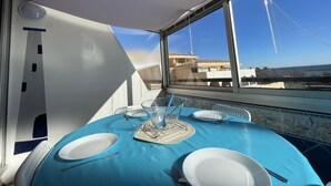 A dining area with a blue tablecloth, white plates, and cutlery. A window reveals a view of buildings and a clear sky.