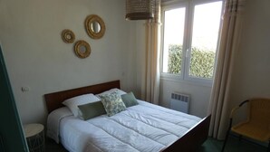 Un letto rifatto con lenzuola bianche e due cuscini, un tavolino rotondo e una sedia in una stanza con un'ampia finestra e un termosifone.