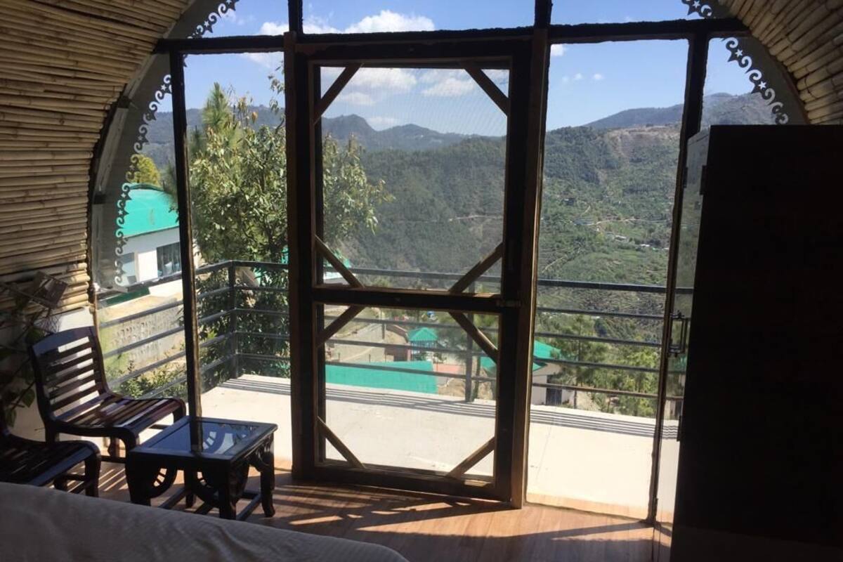 A room with a bed, a wooden bench, and a table. It has a large window with a view of a mountainous landscape.