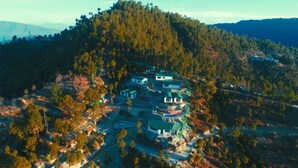 An aerial view of a cluster of buildings on a hillside with dense forest surrounding them. - Dhara Doorway to Inner Peace (Kosiyakutoli)
