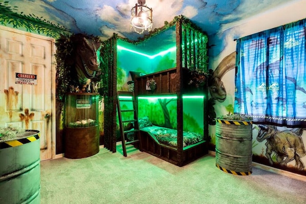 A room with a green dinosaur-themed decor, including a glass enclosure with a dinosaur skeleton, a bed with a green-lit canopy, and a mural of a dinosaur on the wall.