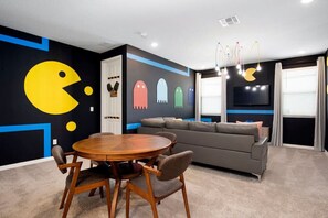 A room with a round wooden table, grey sofa, and a wall with Pac-Man and ghost decorations.