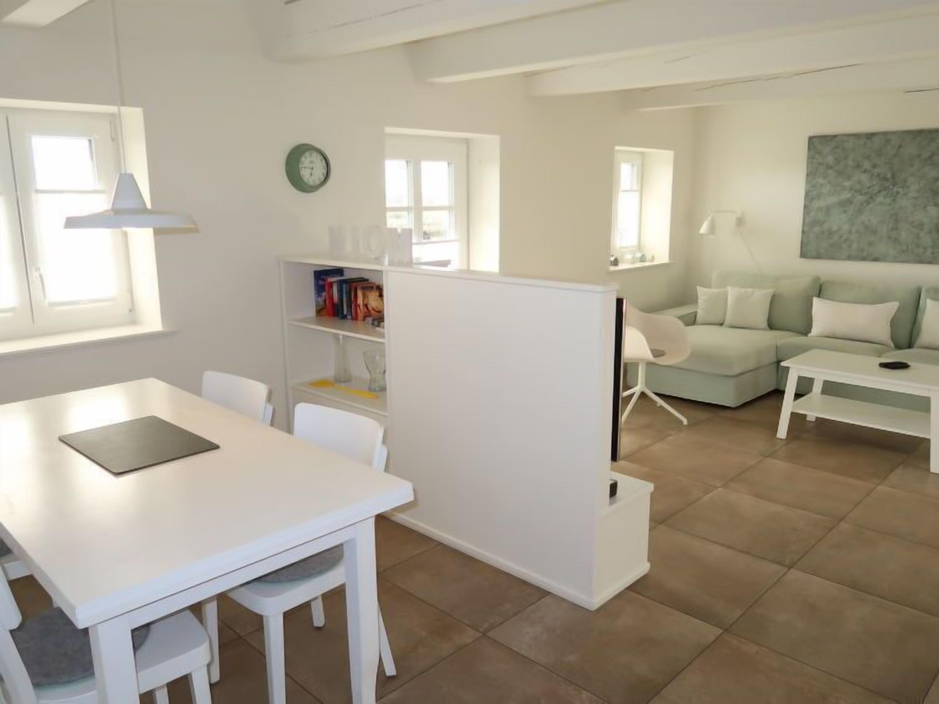 A modern living room with a white sofa set, a dining table with chairs, a bookshelf, and a large window providing natural light.