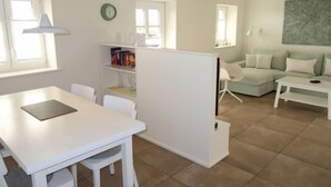 A modern living room with a white sofa set, a dining table with chairs, a bookshelf, and a large window providing natural light.