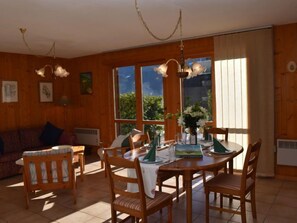 A wooden interior with a dining table set for four, surrounded by chairs. There is a sofa in the background and a large window providing natural light.