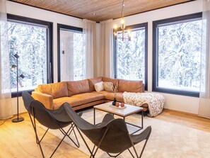 A modern living room with a brown leather sofa, two black chairs, a coffee table, and large windows with a snowy view.