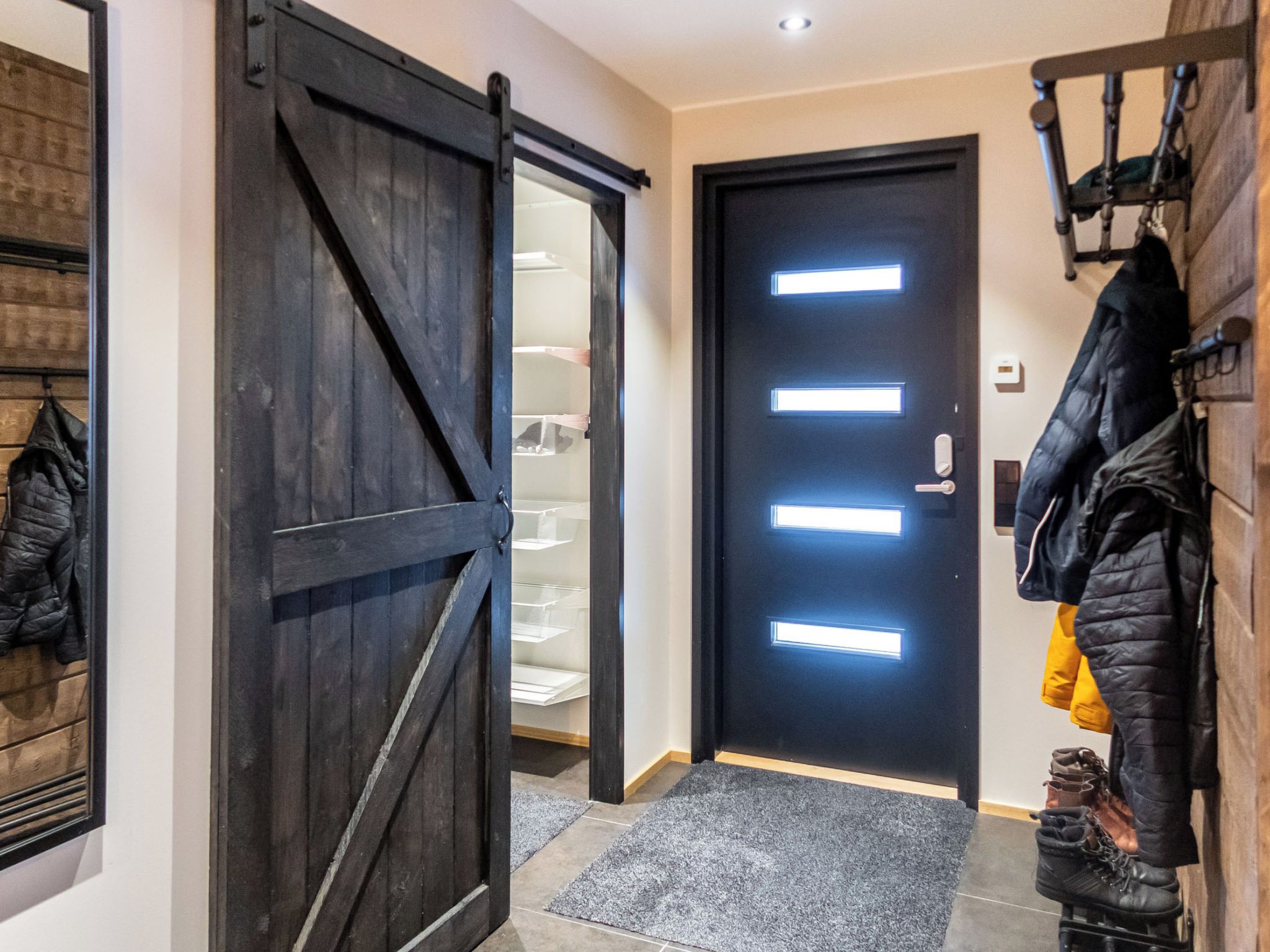 Un pasillo con una puerta de madera oscura, un perchero con chaquetas y un zapatero con botas.