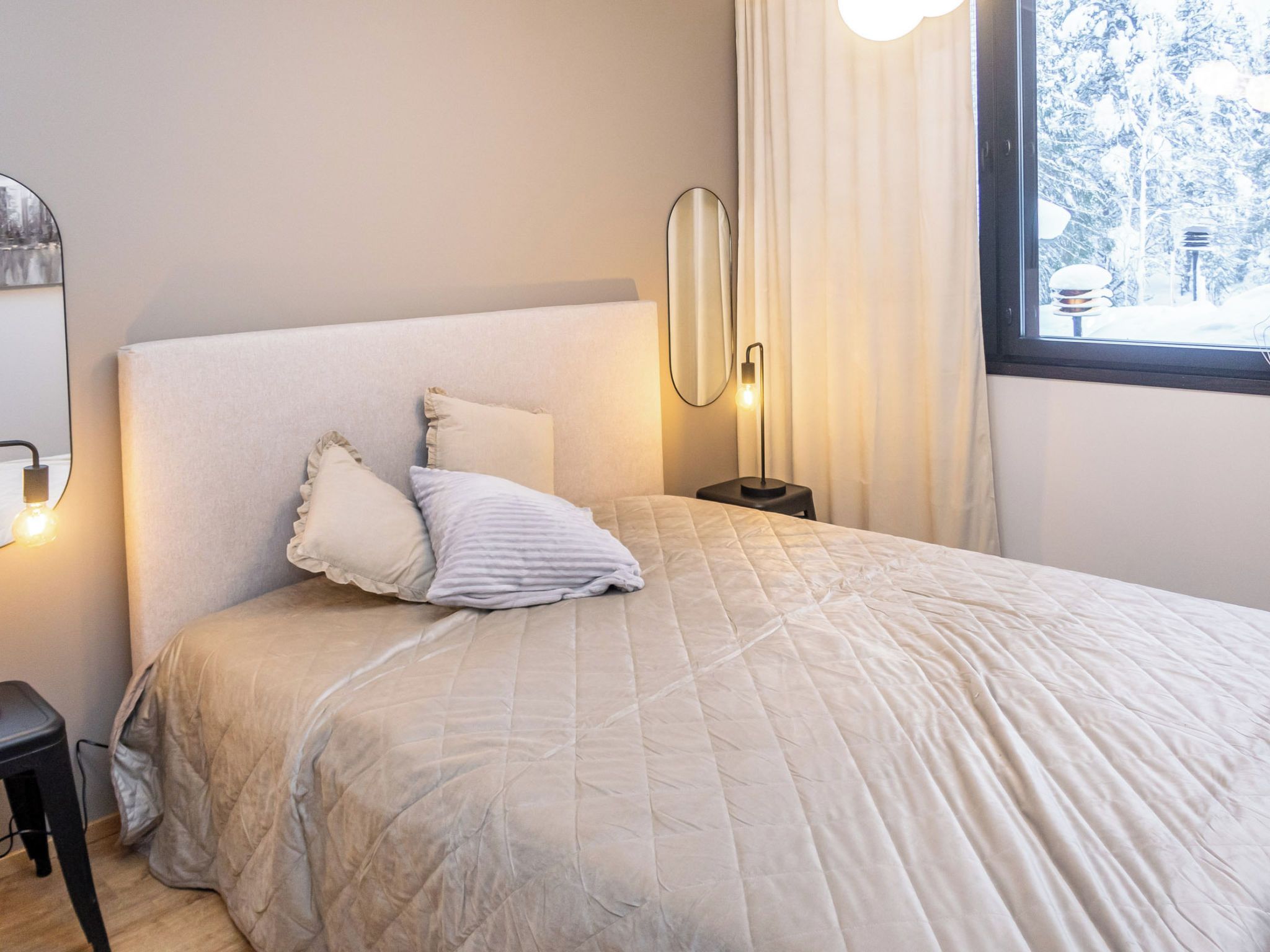 Una cama bien hecha con cubrecama acolchado, una lámpara de noche y una ventana con cortinas.
