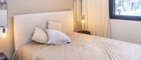 A neatly made bed with a quilted cover, a bedside lamp, and a window with curtains.