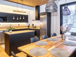 A modern kitchen with a central island, a dining area with a wooden table and chairs, and a view of a snowy landscape through a window. - Vaaranhelmi by Interhome (Kuusamo)