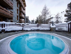 Een bubbelbad omgeven door een stenen gebouw met houten balkons, een met sneeuw bedekte afrastering en bomen op de achtergrond.
