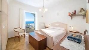 A bedroom with a bed, a wooden nightstand, a desk, a chair, and a view of the sea through an open door. - Raphael House, terrace with solarium and sea view (Conca dei Marini)