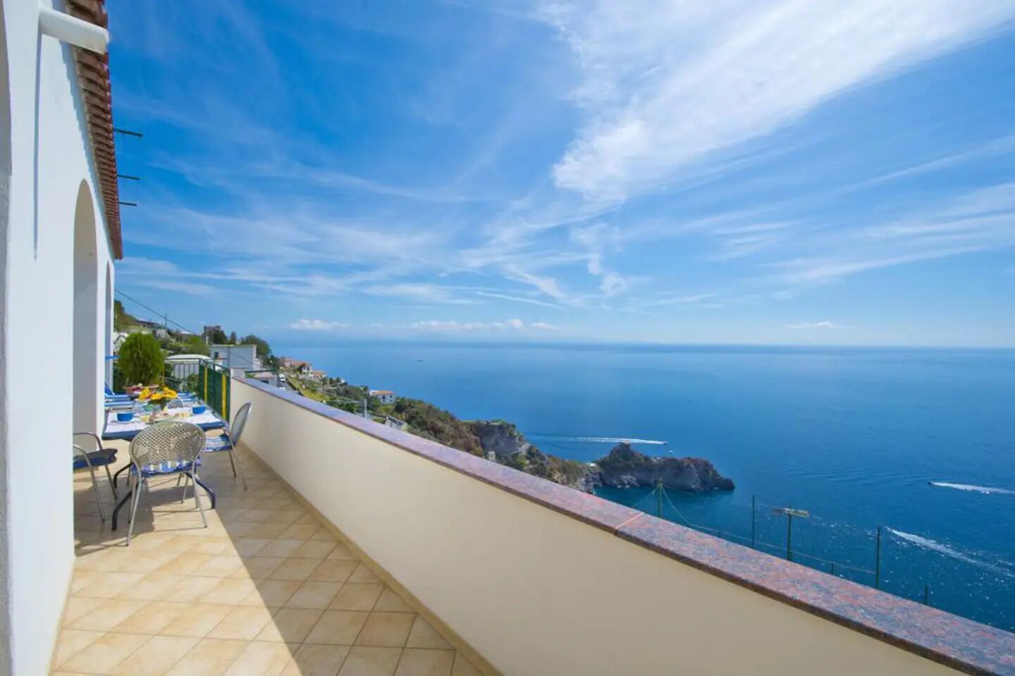 A balcony with a view of the sea, a table with chairs, and a building with a white facade.